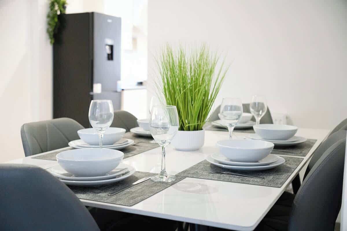 A dining table set with white plates, bowls, and wine glasses, with a potted plant in the center.