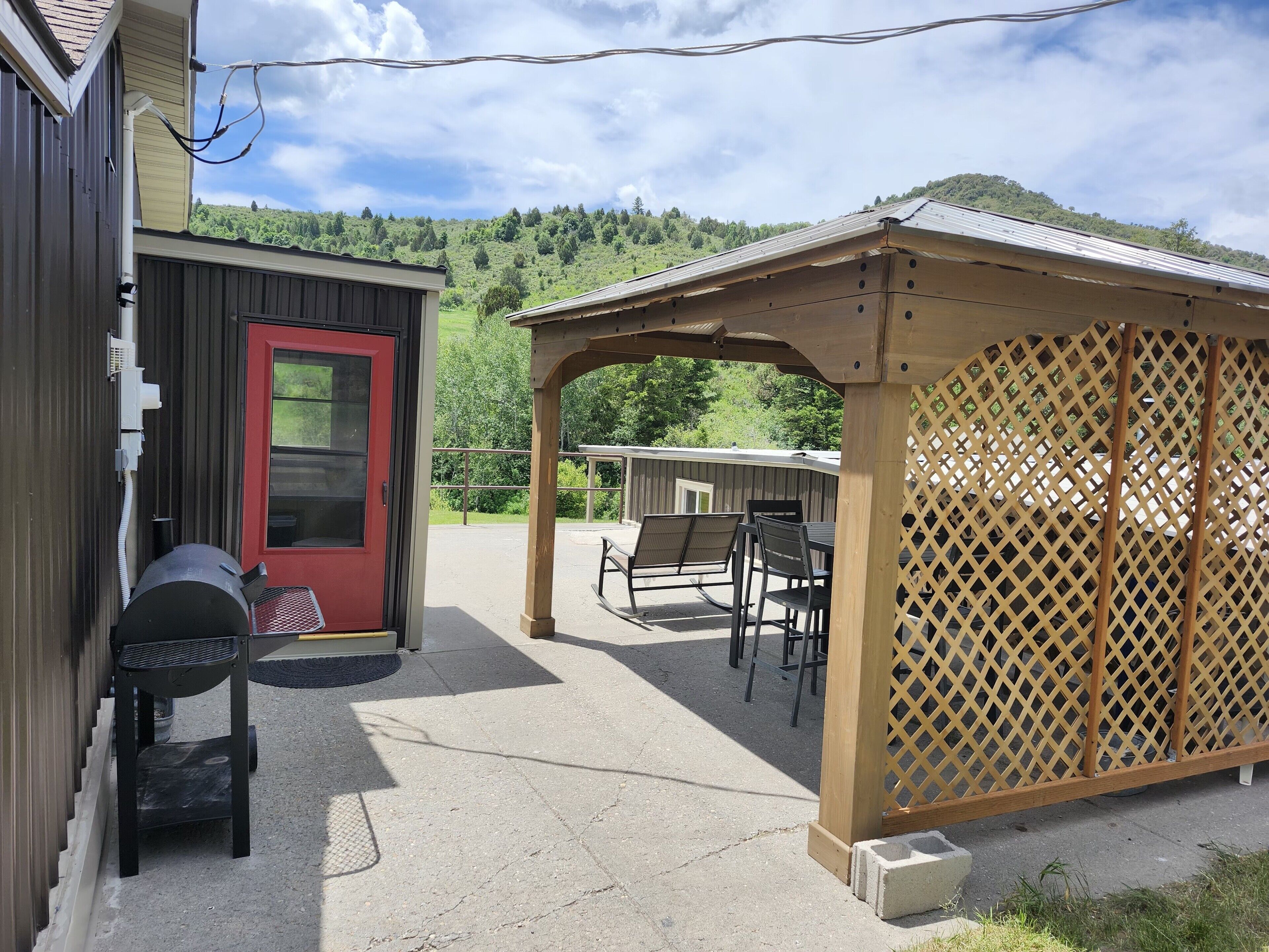 Un espacio de patio cubierto con mobiliario exterior, una parrilla y vistas a una ladera verde.