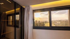 Una habitación con un gran ventanal, cortinas y vistas a un paisaje urbano al atardecer.