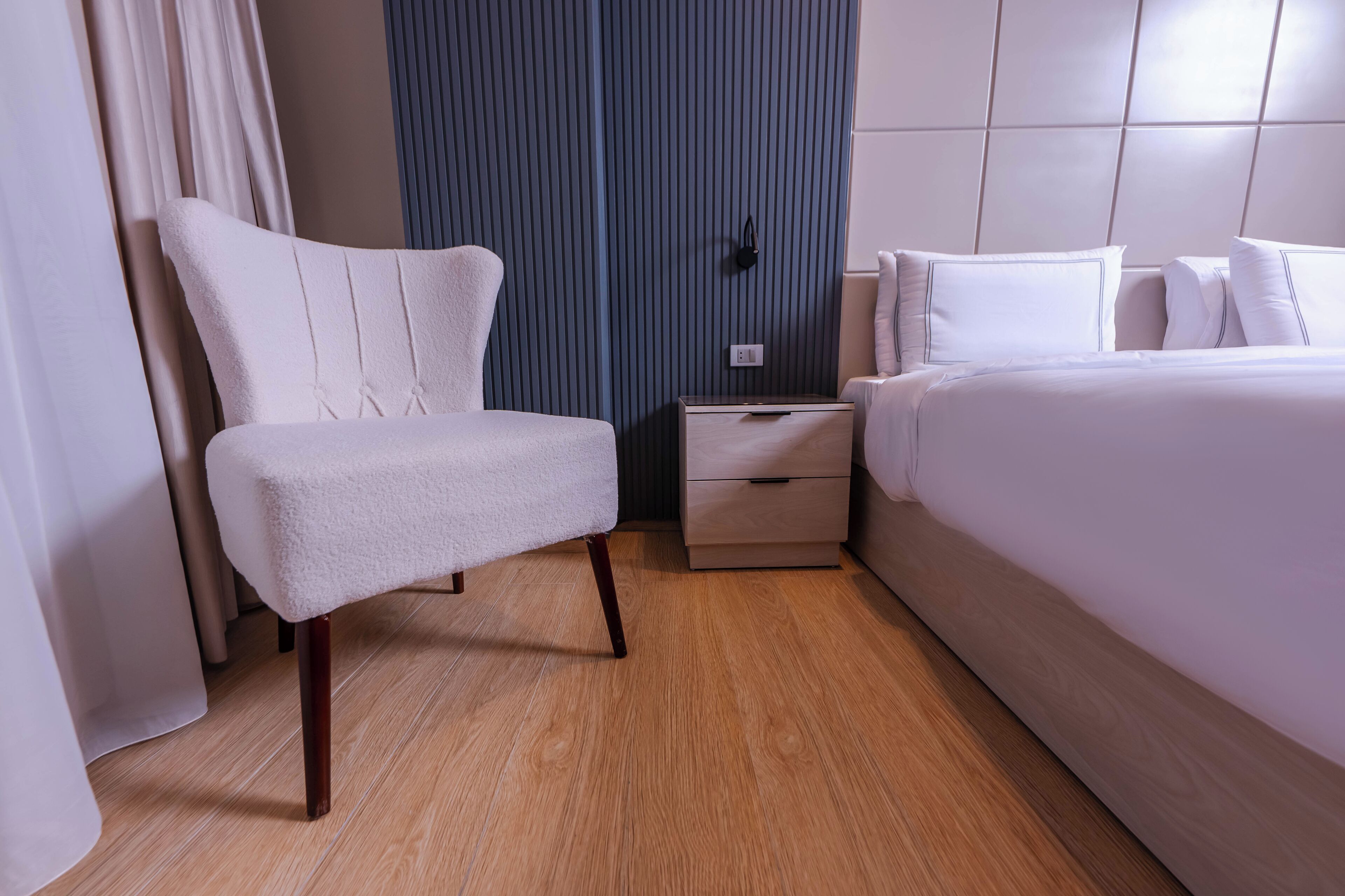 Ett modernt hotellrum med en stor säng, en vit fåtölj, ett träbord som sängbord och en blå accentvägg.