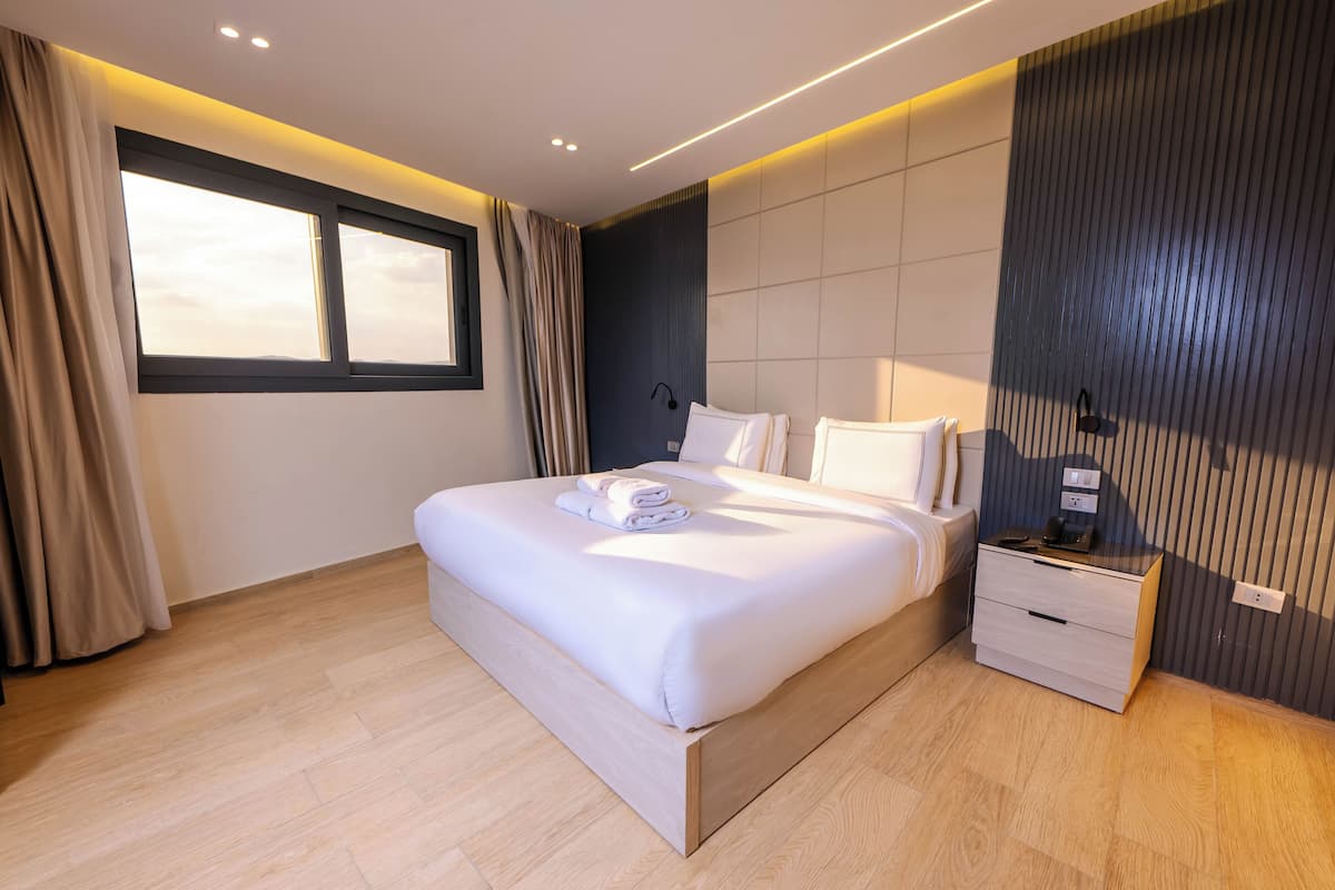 Une chambre moderne avec un grand lit, des tables de chevet, une petite table et une vue sur le ciel par la fenêtre.