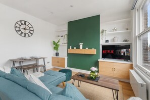 Ein modernes Wohnzimmer mit grüner Akzentwand, einer Wanduhr, einem Essbereich mit Tisch und Bänken sowie einem Fernseher, der 'NETFLIX' anzeigt.