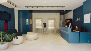 A modern reception area with a blue counter, two elevators, a red door, and a seating area with white poufs. - Apê Santos Gonzaga (Santos)