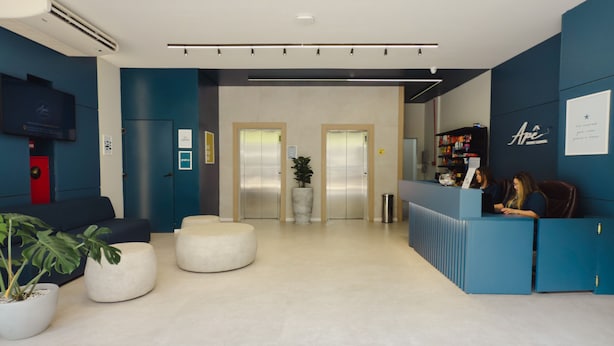 A modern reception area with a blue counter, two elevators, a red door, and a seating area with white poufs. - Apê Santos Gonzaga (Santos)