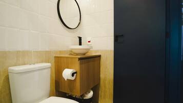 Un baño con inodoro, lavamanos con gabinete de madera, rollo de papel higiénico y una puerta oscura.