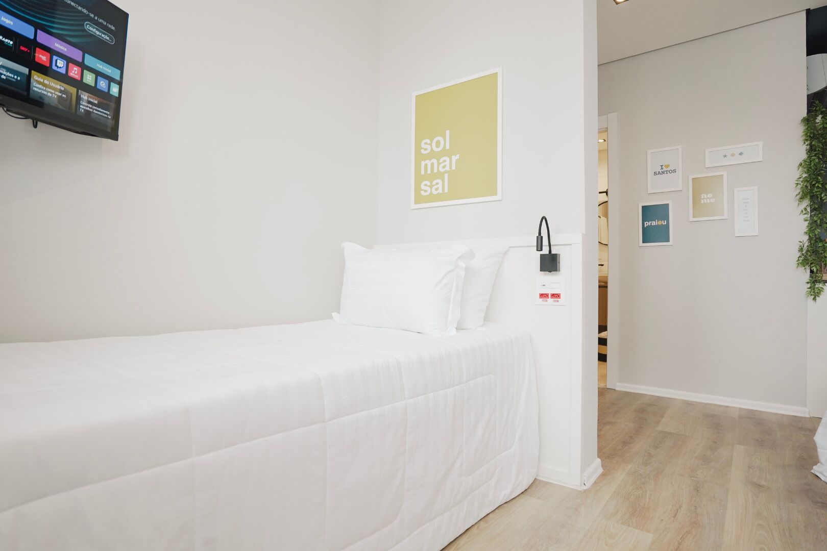 Habitación de hotel con cama, televisor montado en la pared, un cartel que dice "sol mars sal" y una planta pequeña a la derecha.