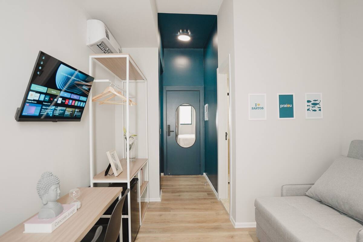 Ein modernes Hotelzimmer mit einem Holztisch, einem grauen Sofa, einem Fernseher, der eine digitale Benutzeroberfläche zeigt, einem Kleiderschrank und einem Badezimmer mit Spiegel.