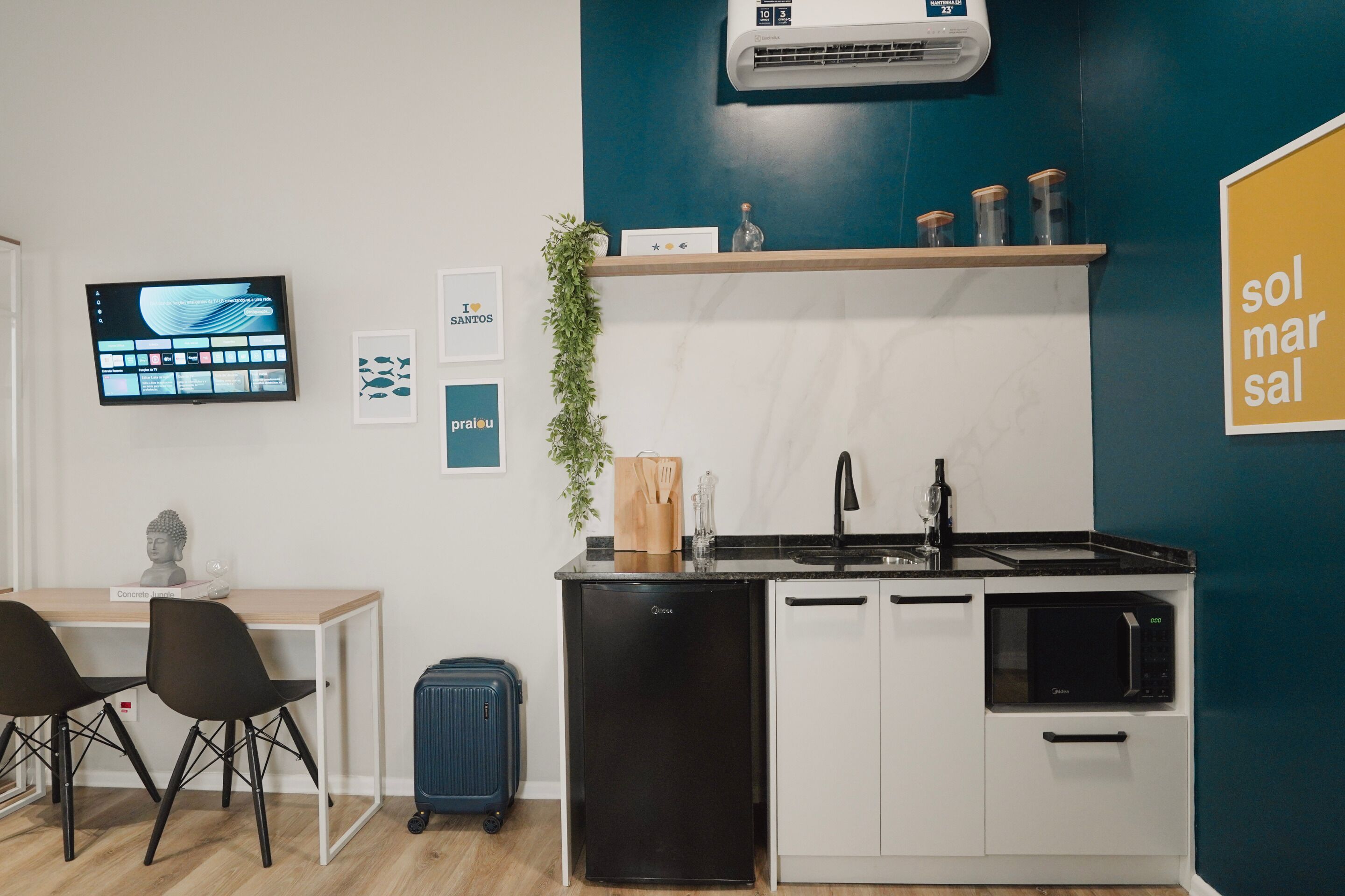 A compact kitchenette with a black refrigerator, white cabinets, a microwave, and a sink. There's a small table with two chairs and a blue suitcase. A flat-screen TV is mounted on the wall, and there are framed pictures and a potted plant.