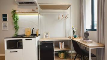 A compact kitchenette with a microwave, refrigerator, and a small sink. There is a wooden desk with a chair, a small shelf with books, and a potted plant.