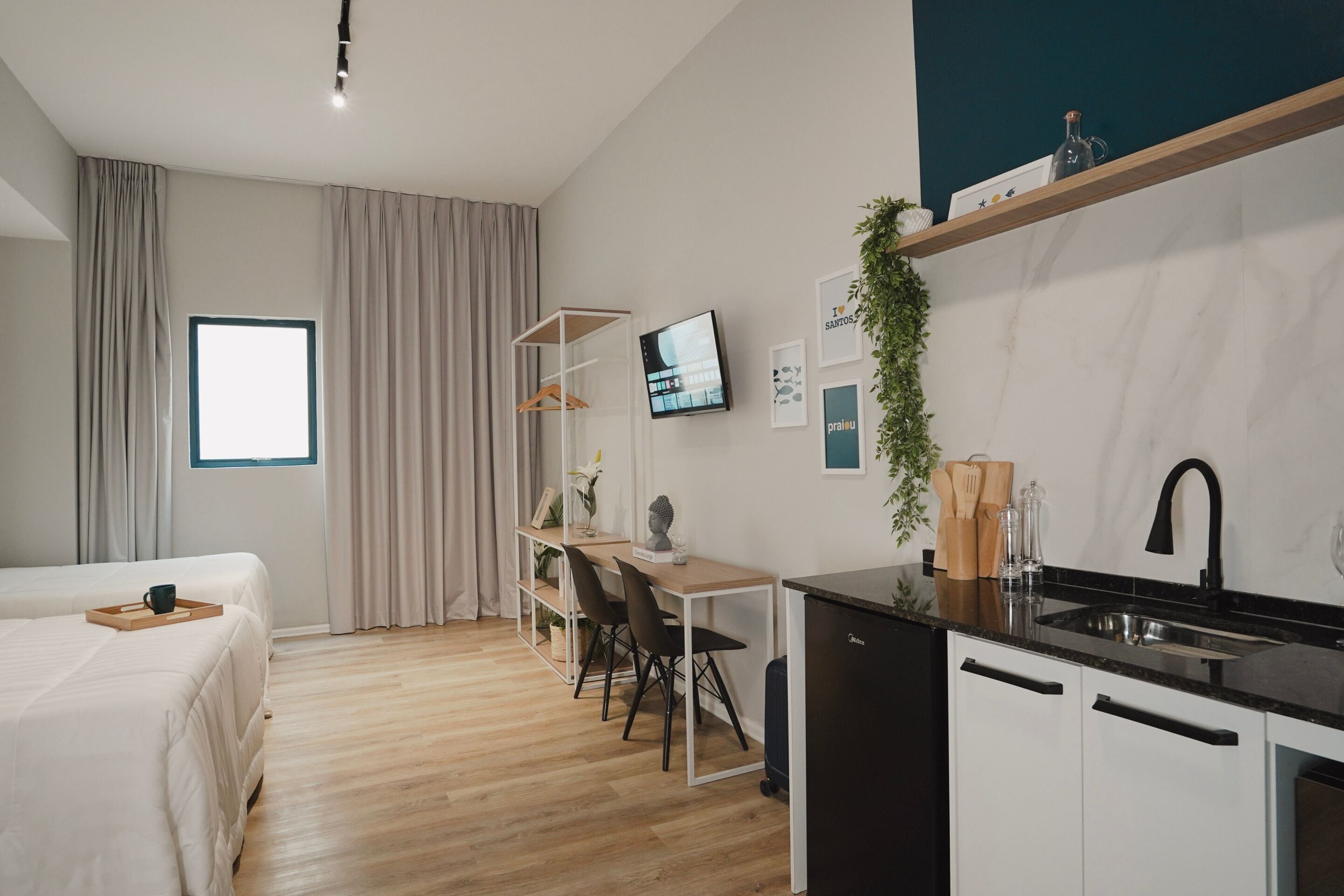 A modern hotel room with a bed, a desk, a TV, a kitchen area, and a window.