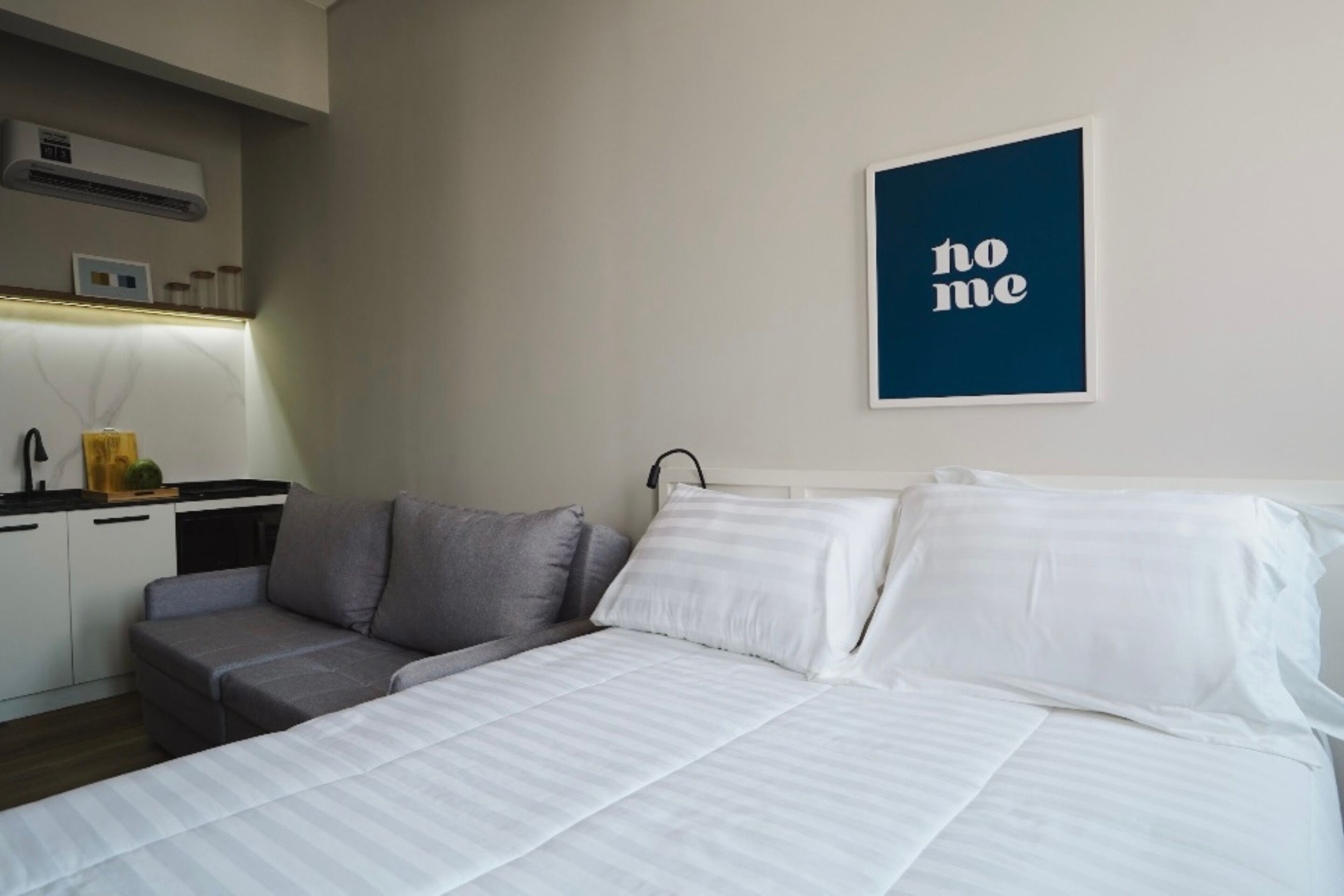 A modern hotel room with a neatly made bed, a gray sofa, and a small kitchenette in the background.