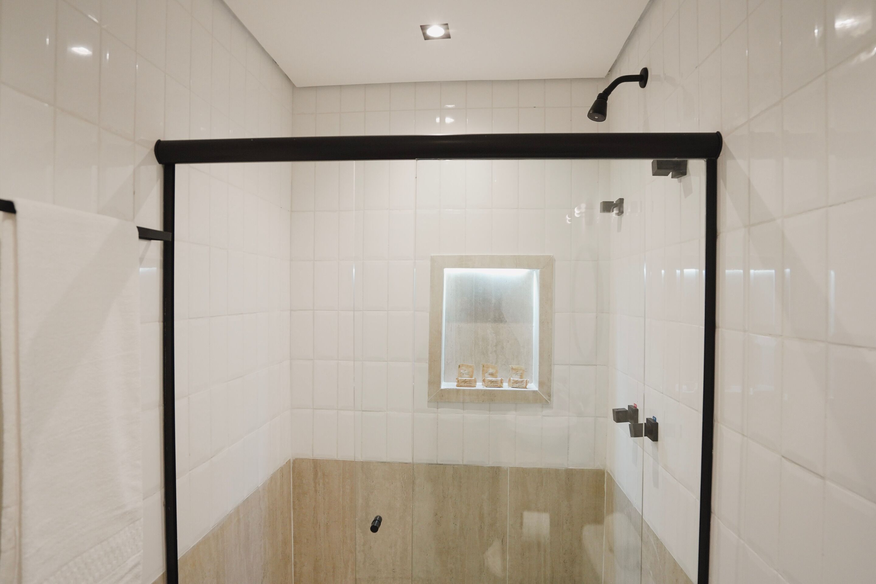 A modern bathroom with white tiles, a glass shower enclosure, a small window with a mirror, and a light fixture on the ceiling.
