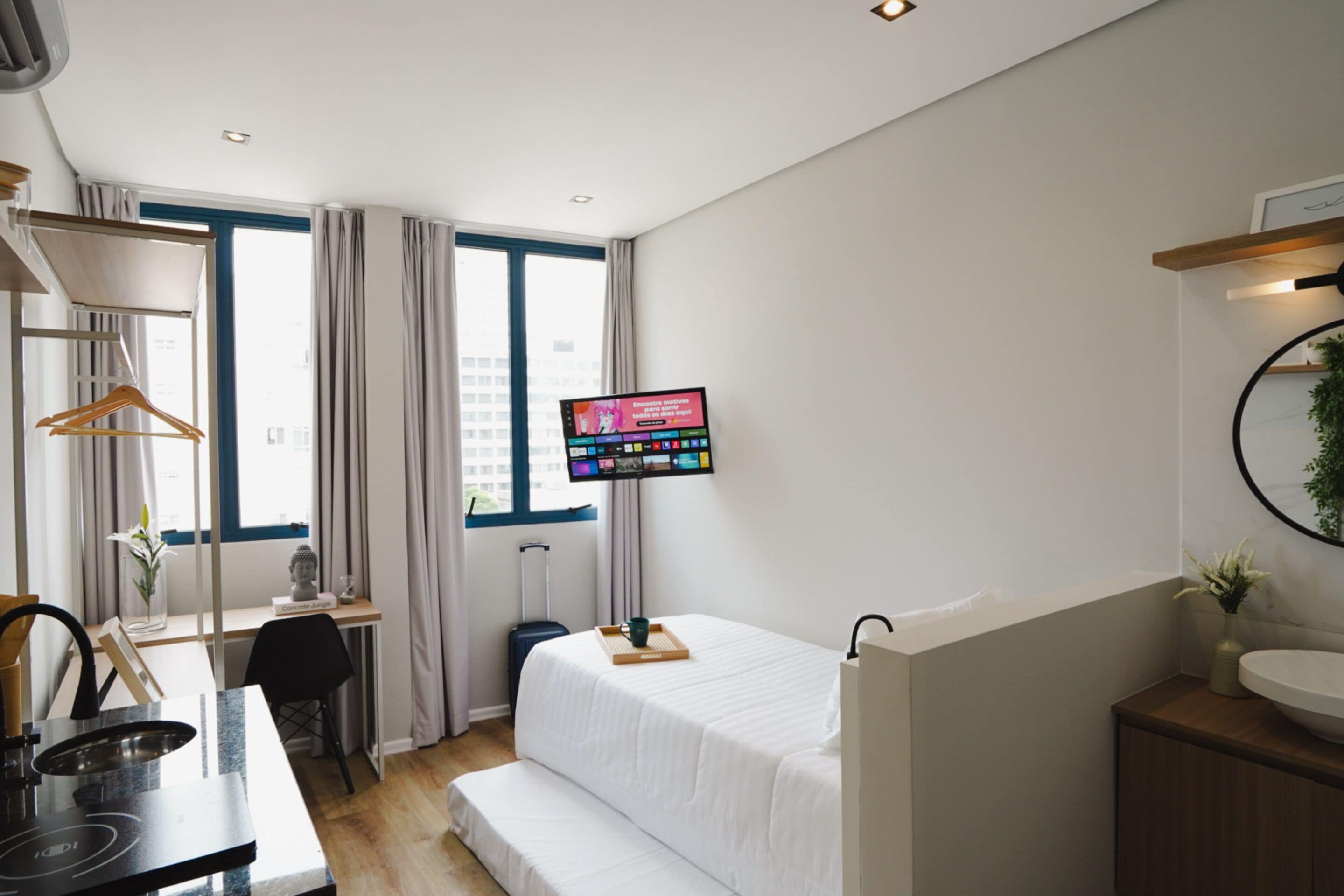A hotel room with a bed, a desk with a computer, a TV mounted on the wall, and a window with curtains.