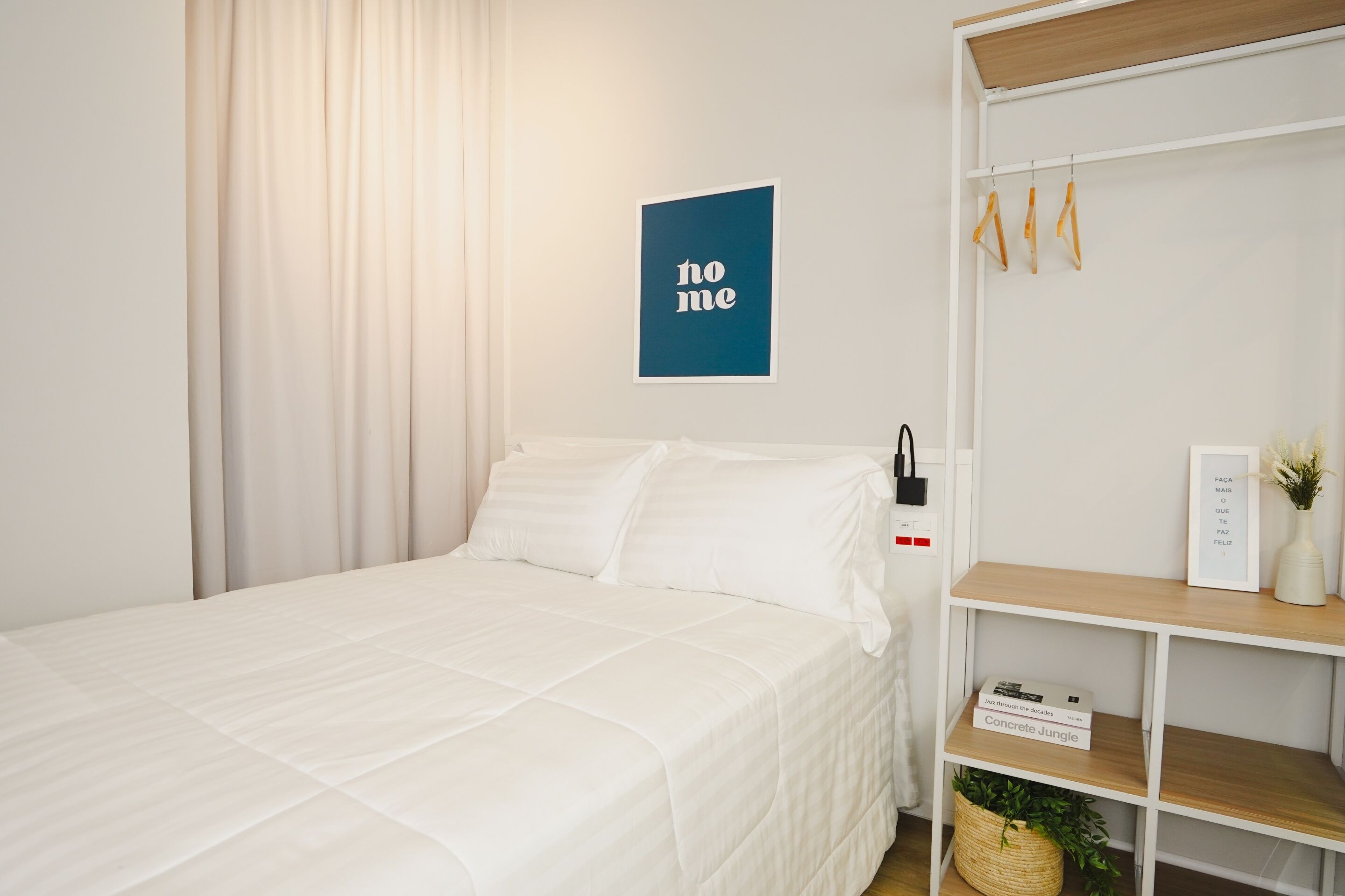 A neatly made bed with white bedding, a blue wall art piece with the word "home", a white wardrobe with hanging items, and a small shelf with a plant and a book.