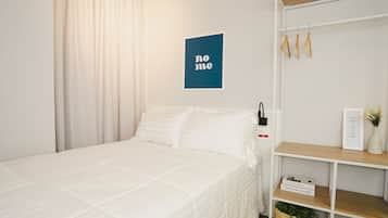 A neatly made bed with white bedding, a blue wall art piece with the word "home", a white wardrobe with hanging items, and a small shelf with a plant and a book.
