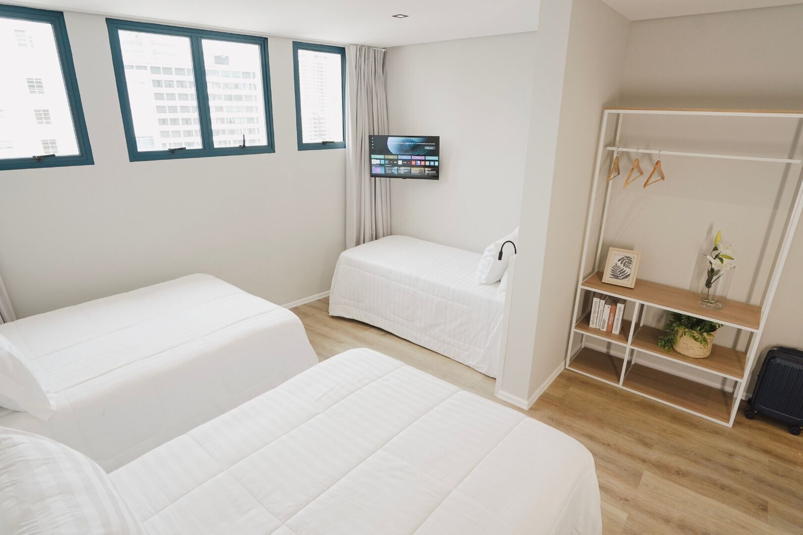 A hotel room with two beds, a TV mounted on the wall, a wardrobe, and a view of buildings through the windows.