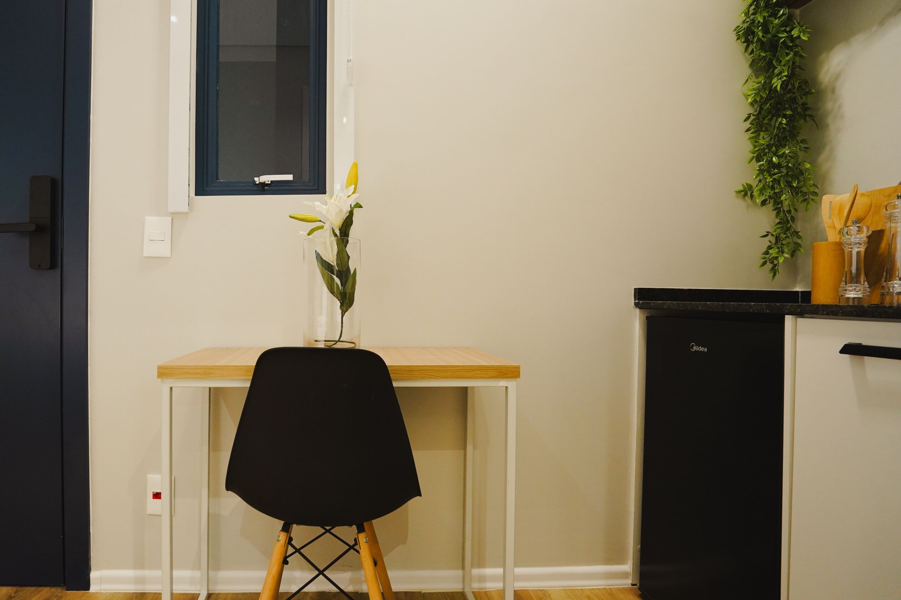 Une petite cuisine avec une table en bois, une chaise noire, un mur blanc, une fenêtre, un lave-vaisselle noir et une plante verte.