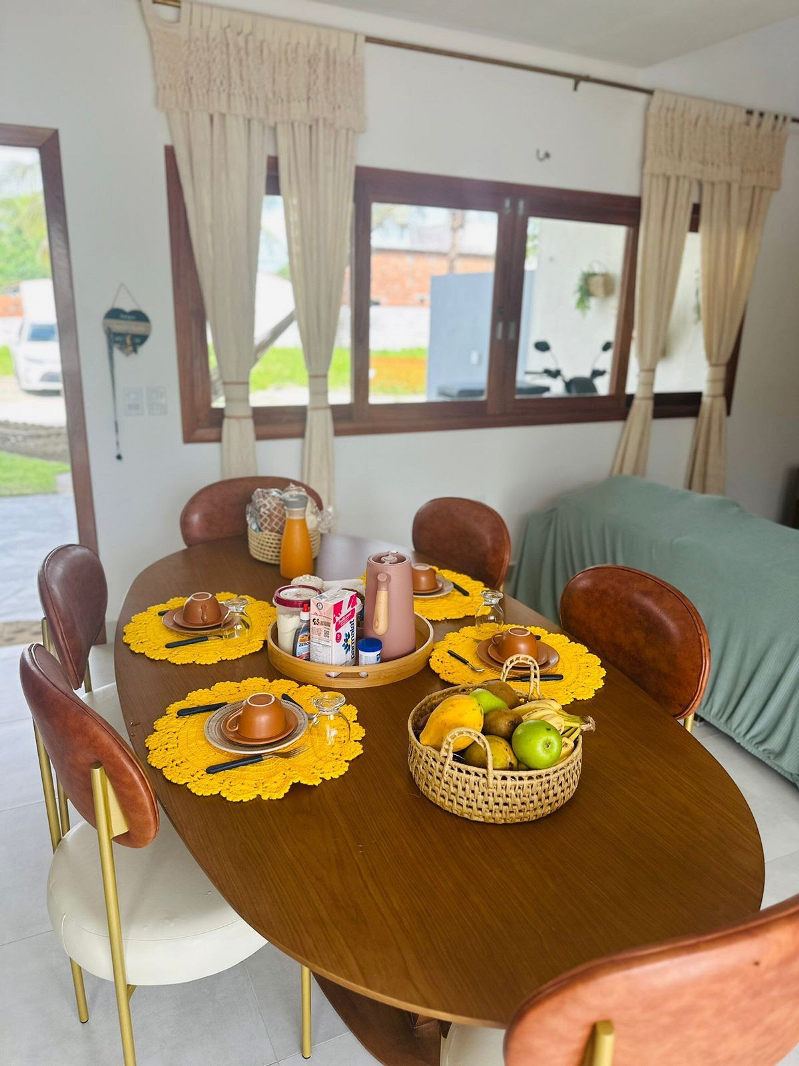 Un coin repas avec une table en bois dressée pour quatre personnes, agrémentée de sets de table jaunes, d’un panier de fruits et de divers condiments.
