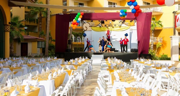 A banquet hall with tables set for dining, a stage with performers, and a yellow building facade.