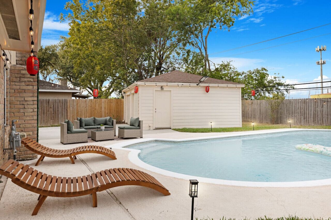 Un jardin avec une piscine, des chaises longues en bois, un patio couvert et une cour clôturée.