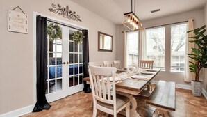 Uma sala de jantar com uma mesa de madeira, cadeiras brancas e um candelabro. Há uma janela com persianas e um vaso de planta.