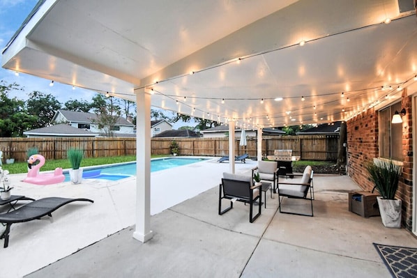 A covered patio area with string lights, a pool, lounge chairs, and a barbecue grill.