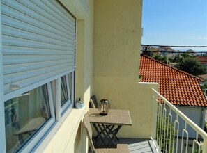 Een balkon met een tafel en stoelen, een raam met zonwering, en een uitzicht op een gebouw met rode dakpannen op de achtergrond.
