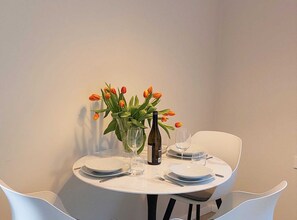 Een ronde eettafel met witte borden, bestek en een fles wijn. Er zijn twee glazen en een vaas met oranje tulpjes. Witte stoelen staan rond de tafel.