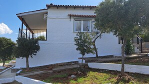 A white building with a red-tiled roof, a balcony, and a small window. There are trees and a stone wall in the foreground. - House by the sea (Salamina)