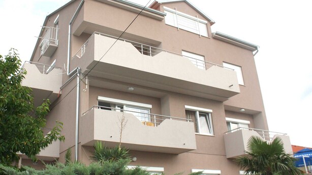 A multi-story residential building with balconies, windows, and a small tree in the foreground.
