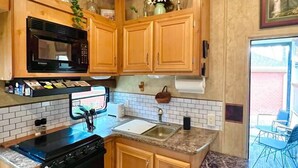 A compact kitchen with wooden cabinets, a microwave, a stove, and a sink. - The Glamper Spacious 5th Wheel (Temple)