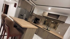 A modern kitchen with a marble floor, wooden bar stools, a built-in oven, and a stainless steel refrigerator. - 3-ROOM APARTMENT WITH BALCONY TERRACE GARDEN SWIMMING POOL (Abidjan)