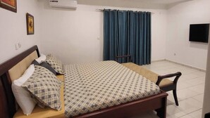 A bedroom with a bed, a chair, a TV, and an air conditioning unit. - 3-ROOM APARTMENT WITH BALCONY TERRACE GARDEN SWIMMING POOL (Abidjan)