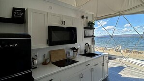 A modern kitchen with white cabinetry and a black stove, under a large white tent structure with a view of the beach. - Dome 2 · Luxury Lakeside Dome With Hot Tub (St. George`s Channel)