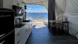 A compact kitchen with modern appliances, a dining area with a table and chairs, and a view of the sea through a large tented window. - Dome 2 · Luxury Lakeside Dome With Hot Tub (St. George`s Channel)