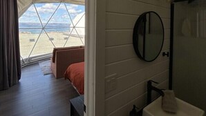 Una habitación con un ventanal grande con vista a una playa, una cama, una mesita y un baño con espejo y lavamanos.