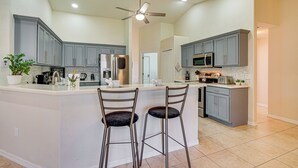 A modern kitchen with grey cabinets, a central island with two bar stools, stainless steel appliances, and a ceiling fan. - 4 Mi to Beach: Home w/ Patio in Cape Coral! (Cape Coral)