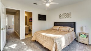 Un dormitorio con cama, mesita de noche, ventilador de techo, televisor y una puerta que da a otra habitación.