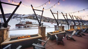 A terrace with outdoor seating and fire pits, overlooking a snowy mountain area.