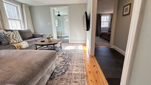 A living room with a sofa, coffee table, and a rug. There is a TV mounted on the wall and a door leading to another room. - 12 Mins from Beach - A Quiet, Immaculate space (Norwell)