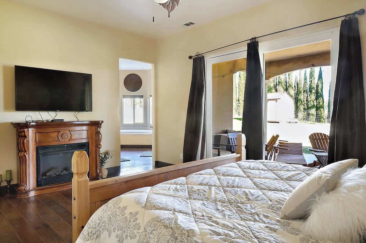 A bedroom with a large bed, a fireplace, a television, and a view of the outdoors.