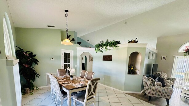 Un coin repas avec une table dressée pour quatre personnes, entourée de chaises blanches. Un lustre se trouve au-dessus de la table, et une plante est visible à l’arrière-plan.