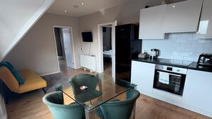 Een moderne keuken met witte kastjes, een zwarte koelkast en een inbouwoven. Er is een eethoek met een glazen tafel en twee groene stoelen.