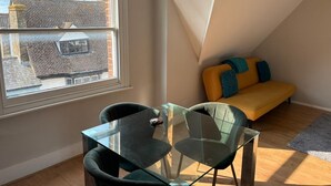Een kleine, moderne kamer met een glazen tafel en stoelen, een gele bank en een groot raam met uitzicht op een gebouw van baksteen.