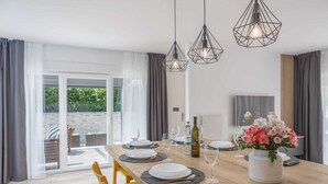 A dining area with a wooden table set for four, three pendant lights, a vase of flowers, and a bottle of wine. - Apartment BARBARA with private pool (Malinska)