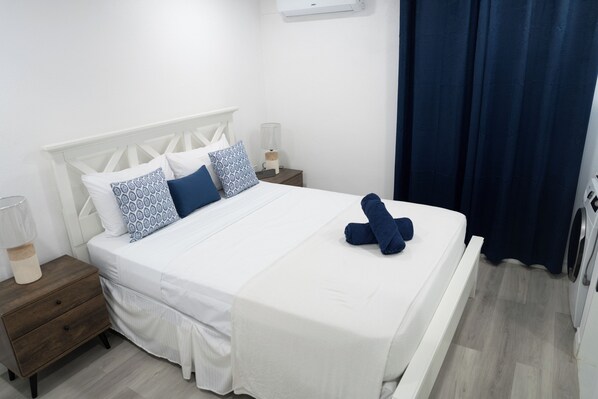 A neatly made bed with white bedding and blue pillows, a wooden nightstand with a lamp, and a dark blue curtain partially drawn.