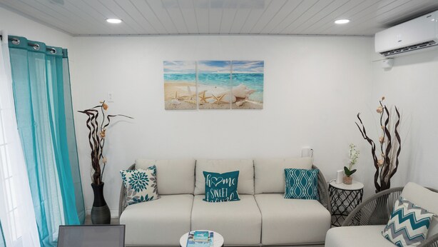 A modern living room with a beige sofa, teal cushions, a coffee table, and a wall-mounted painting of a beach scene. -  Cosy one bedroom condo in Holetown, St. James with pool access!! (Holetown)