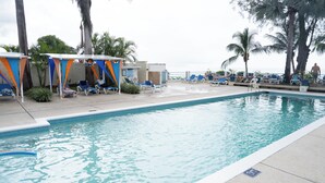 A swimming pool with lounge chairs and umbrellas, surrounded by palm trees and a view of the ocean. -  Cosy one bedroom condo in Holetown, St. James with pool access!! (Holetown)