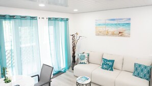 A modern living room with a white sofa, teal cushions, a coffee table, and a painting of a beach scene. -  Cosy one bedroom condo in Holetown, St. James with pool access!! (Holetown)