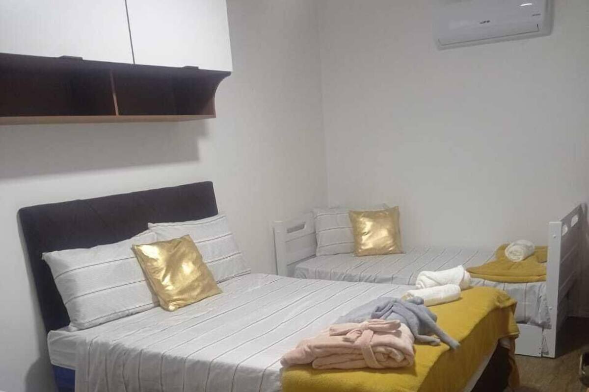 A dormitory room with three beds, each with a different colored bedspread. There is a white air conditioning unit on the wall, and a wooden table in the corner.